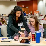 two veterinary students work together at a desk in a room full of trainees, one student stands to assist the other 