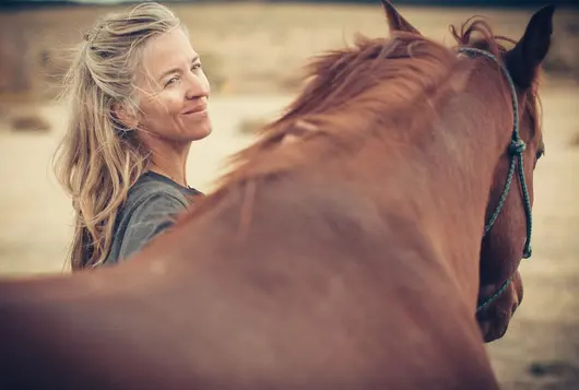 a woman stands next to a brown horse in the wind smiling