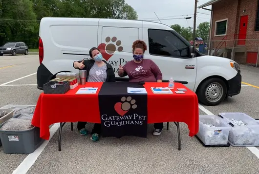 two workers sit at a table in front of a pet food distribution van giving the thumbs up sign