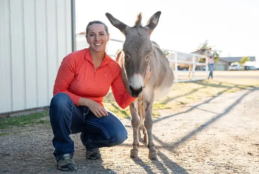 a woman wearing an orange top kneels next to a tan donkey near a barn structure