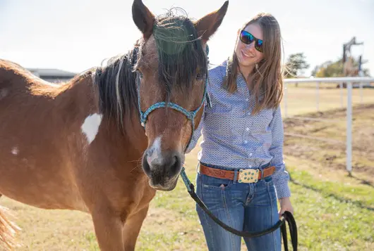 a woman wearing a blue shirt stands outside looking happily at a brown horse